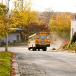 A yellow school bus on a village road during a crisp autumn day, surrounded by colorful trees.