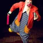A vibrant clown in striped costume joyfully performs on a wooden stage, exuding happiness and humor.